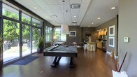 Located at 250 Brodbeat Ln in Columbus, Ohio, this single-floor amenity space is thoughtfully designed to provide a welcoming and versatile environment for recreation and socializing. The open-concept layout is anchored by a full-size billiards table positioned beneath contemporary pendant lighting, creating a natural focal point for the room. Rich, dark hardwood flooring runs throughout, complementing the warm gray tones of the accent walls and lending a polished, modern aesthetic. A wall of floor-to-ceiling windows along one side floods the space with abundant natural light and offers views of lush greenery outside, enhancing the overall ambiance with a connection to the outdoors. Potted plants add to the organic feel of the interior. Toward the back of the space, a well-equipped kitchenette features light wood cabinetry, stainless steel appliances, and a counter with bar-height seating — ideal for casual gatherings. A recessed ceiling with flush lighting and a drop-panel ceiling section provide layered illumination throughout. Decorative wall art and signage contribute to the space's urban, contemporary character. The combination of recreational amenities, natural light, and functional design makes this a standout communal space.