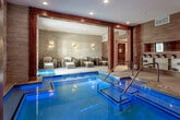 The Donovan, located at 837 NW Donovan Rd in Lee's Summit, Missouri, is a sophisticated spa and aquatic wellness facility designed with a harmonious blend of natural materials and modern elegance. The space is anchored by a luminous indoor pool with glowing blue-lit waters, accessible via stainless steel handrails and entry steps, and framed by striking exposed timber beams that lend a warm, lodge-inspired character to the interior. The "THE DONOVAN" branding is prominently displayed along the textured stone wall, reinforcing the facility's upscale identity. Surrounding the pool, light travertine tile flooring extends into a generously appointed lounge area furnished with woven rattan chairs and matching ottomans, each fitted with plush white cushions and accented with striped throw pillows. Warm sconce lighting mounted against the stacked stone feature walls creates an intimate, calming ambiance throughout the space. Warm-toned wood paneling complements the natural stone textures, while recessed ceiling lighting ensures the entire area is well-illuminated. An adjoining relaxation alcove offers additional seating away from the pool, ideal for pre- or post-treatment rest. The overall design strikes a balance between spa tranquility and resort-style comfort, making The Donovan a refined destination for wellness and relaxation.