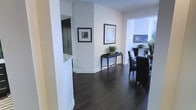 This well-appointed single-floor space exudes a clean, contemporary aesthetic defined by a thoughtful combination of materials and design choices. Rich, dark hardwood flooring extends throughout, providing a striking contrast against the bright white walls and baseboards that lend the space an open, airy feel.

The entryway sets an elegant tone, featuring a sleek black console table adorned with a decorative tray and a potted plant, flanked by framed artwork that adds a curated, sophisticated touch. A large mirror reflects light and enhances the sense of depth along one wall, making the entry feel both welcoming and spacious.

Moving further into the space, a dining area comes into view, furnished with dark upholstered chairs arranged around a glass-top table. Lush greenery in the form of indoor plants adds a natural warmth that softens the overall modern aesthetic. A flat-screen television is visible in the background, hinting at a connected living area beyond.

The seamless flow between areas, consistent flooring, and neutral color palette create a cohesive environment throughout. The balance of dark furnishings against light walls gives the space a timeless elegance, while the carefully selected décor and greenery infuse it with personality and life. Overall, this is a refined and functional space designed for comfortable, stylish living.