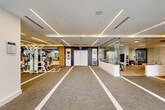 Located at 101 E Camino Real in Boca Raton, Florida, this upscale fitness center offers a generously proportioned, single-floor layout designed to support a comprehensive range of workout activities. The space is defined by its sleek, contemporary architecture, featuring linear recessed lighting embedded in tiered ceilings that cast a clean, even illumination across the entire floor plan.

The main workout area is divided into distinct zones. A robust strength training section houses a wide selection of cable machines, multi-functional weight stations, free weights, and benches, all arranged to maximize accessibility and flow. Rubber floor mats provide safety and cushioning in lifting areas, while neutral-toned carpet with bold white stripe detailing guides movement throughout the space.

A dedicated cardio zone lines the perimeter near floor-to-ceiling glass windows, offering a row of treadmills and other cardio equipment with direct views of the outdoor pool terrace and surrounding Florida landscape. Glass-paneled walls allow natural light to penetrate deep into the facility.

A separate group exercise studio features light wood flooring, full-length mirrors, wall-mounted flat-screen displays, and a dark accent feature wall, creating an inviting environment for classes and personal training. The overall ambiance is modern, bright, and professionally appointed, reflecting a high-end fitness facility.
