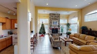This single-floor space is designed as an open, communal gathering area that combines comfort and functionality. The centerpiece of the main lounge is a floor-to-ceiling stacked stone fireplace, which anchors the room and creates a warm, inviting focal point. Surrounding it, generous seating is arranged thoughtfully, including tan leather sofas, patterned armchairs, and a wooden coffee table set atop an area rug — all contributing to a relaxed, residential atmosphere.

The soaring ceilings feature recessed lighting and are complemented by warm yellow accent walls that contrast with crisp white trim and soffits, lending a bright, cheerful character throughout. Oversized windows with white blinds line multiple walls, allowing natural light to fill the space and offering views of the surrounding exterior.

A secondary seating area near the entrance provides additional intimate conversation space, while a built-in counter with bar-height seating offers a casual spot for quick use. Adjacent to this area is a compact, well-appointed kitchen nook equipped with wood cabinetry, a double sink, electric range, microwave, and ample counter space. Light tile flooring runs consistently throughout, tying all areas of the space together cohesively.