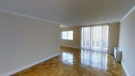 This single-floor space presents a clean, open layout with notable architectural details and quality finishes throughout. The living area is characterized by its polished parquet wood flooring, which features a classic block pattern that reflects light and adds warmth and visual interest underfoot. Crown moulding runs along the perimeter of the ceiling, providing a refined finishing touch that elevates the overall aesthetic of the room.

Natural light is a defining feature of this space. A large sliding glass door fitted with full-length vertical blinds occupies a substantial portion of one wall, allowing for generous light intake and providing access to what appears to be an outdoor area or balcony. A secondary window on the adjacent wall further enhances the brightness of the room, ensuring even illumination throughout the day.

The walls are finished in a neutral, warm beige tone that complements the flooring and contributes to a calm, welcoming atmosphere. White baseboards provide crisp contrast along the floor line. The space is open and unobstructed, offering flexibility for a variety of furniture arrangements and uses.