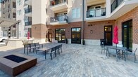 Located at 76 N Center St in the heart of downtown Hickory, North Carolina, this single-level outdoor courtyard space is set within a mixed-use development characterized by striking red brick facades and modern architectural detailing. The courtyard is paved throughout with variegated stone pavers, creating a polished yet durable surface that anchors the open-air environment.

The space is framed on multiple sides by the surrounding multi-story building, which features large black-framed glass doors and windows at street level, providing direct interior access and allowing natural light to flow freely. Upper-level balconies with black iron railings overlook the courtyard, adding vertical interest and a sense of enclosure without sacrificing openness to the sky.

Furnished with multiple sets of metal tables and chairs, the courtyard is well-suited for outdoor dining, social gatherings, or events. Red market umbrellas provide shade throughout, while a covered pergola structure at one end offers additional shelter. Potted greenery accents the entryways, softening the hard surfaces. The overall ambiance is clean, urban, and inviting, reflecting the character of downtown Hickory's revitalized corridor.