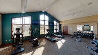 The Bridgewater Apartments fitness center is a well-equipped, single-floor facility designed to support a comprehensive workout experience. The room's most striking architectural feature is its vaulted ceiling, which soars upward and is accentuated by a bold teal accent wall on one side and warm beige tones on the other, creating a visually dynamic and energizing atmosphere. Large windows with wood-trimmed frames line the teal wall, flooding the space with generous natural light and offering views of the surrounding outdoor greenery.

The cardio zone occupies one side of the room and includes multiple treadmills, elliptical trainers, and a rowing machine, several of which are positioned in front of wall-mounted televisions for entertainment during workouts. The flooring in this area features a wood-look surface that transitions to patterned carpet tile in the strength training zone.

The strength training area is comprehensively outfitted with a full rack of dumbbells, an adjustable weight bench, stability balls, medicine balls, and a multi-station cable machine. A large wall mirror enhances the sense of space and allows for form monitoring. Near the entrance, a seating bench and open storage shelving provide practical convenience. The Bridgewater branding is prominently displayed on the wall, reinforcing the facility's identity throughout the space.