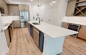 This well-appointed 740-square-foot, one-bedroom, one-bathroom apartment is designed with a modern aesthetic and thoughtful layout across a single floor. The open-concept kitchen and living area serves as the heart of the unit, featuring a large white quartz waterfall island with a black matte faucet and pendant globe lighting overhead. The kitchen is outfitted with flat-front light wood cabinetry, white quartz countertops, and a full suite of stainless steel appliances including a refrigerator, range, and microwave. A built-in wine rack and beverage cooler add a refined touch to the living area. Light wood-look luxury vinyl plank flooring runs seamlessly throughout the unit, complementing the bright white walls and clean trim. The 150-square-foot primary bedroom is bathed in natural light from large windows with roller shades, and includes a ceiling fan with integrated lighting. The spacious bathroom is a standout feature, showcasing a navy blue double vanity with LED-lit mirrors, a glass-enclosed walk-in shower with tile surround, a soaking tub, and large-format tile flooring. A dedicated closet completes this sleek, move-in-ready residence.