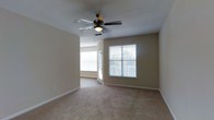 This single-story space presents a clean, functional layout centered around a spacious carpeted room finished in neutral beige tones. The room is fitted with plush wall-to-wall carpeting that adds warmth underfoot, while the smooth, light-colored walls create a bright and airy atmosphere. A ceiling fan with a built-in light fixture is centrally mounted, providing both air circulation and supplemental lighting. Two windows dressed with white horizontal blinds allow generous natural light to fill the space, offering views of the outdoor surroundings. A doorway visible from the main area leads to an adjacent room or space, suggesting a connected, open flow throughout the single floor. White baseboards provide a crisp contrast against the beige walls and carpeting, contributing to a tidy, well-maintained appearance. The overall ambiance is calm and neutral, offering a versatile canvas well-suited for a variety of furnishing styles. The combination of natural light, soft carpeting, and ceiling fan makes this a comfortable and practical living environment.