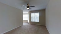 This single-story space presents a clean, functional layout centered around a spacious carpeted room finished in neutral beige tones. The room is fitted with plush wall-to-wall carpeting that adds warmth underfoot, while the smooth, light-colored walls create a bright and airy atmosphere. A ceiling fan with a built-in light fixture is centrally mounted, providing both air circulation and supplemental lighting. Two windows dressed with white horizontal blinds allow generous natural light to fill the space, offering views of the outdoor surroundings. A doorway visible from the main area leads to an adjacent room or space, suggesting a connected, open flow throughout the single floor. White baseboards provide a crisp contrast against the beige walls and carpeting, contributing to a tidy, well-maintained appearance. The overall ambiance is calm and neutral, offering a versatile canvas well-suited for a variety of furnishing styles. The combination of natural light, soft carpeting, and ceiling fan makes this a comfortable and practical living environment.