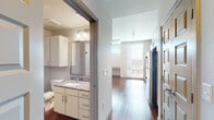 This single-floor unit at 1232 Delaware St in Denver, CO offers a well-designed layout with contemporary finishes throughout. The kitchen is a standout feature, equipped with stainless steel appliances including a refrigerator, range, microwave, and dishwasher. White shaker-style cabinetry pairs with granite-look countertops and a white subway tile backsplash to create a clean, modern aesthetic. A large floor-to-ceiling window floods the kitchen and adjacent living area with natural light while offering views of the surrounding Denver neighborhood.

Wood-style flooring runs throughout the unit, adding warmth and continuity to the space. The living area is generously proportioned, featuring a ceiling fan with a track lighting fixture and neutral white walls that provide a versatile backdrop.

The bathroom is well-appointed with white cabinetry, granite-look countertops, a large vanity mirror, and a shower-tub combination. A sliding barn-style door provides a space-efficient entry to the bathroom. Upper wall cabinets offer additional storage.

The hallway connects the living, kitchen, and bathroom areas in a practical and efficient floor plan. The unit's neutral color palette, modern hardware, and quality finishes contribute to a cohesive and contemporary overall ambiance.