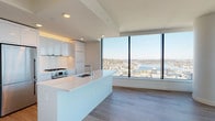 This sophisticated high-rise unit offers a clean, modern aesthetic defined by an open-concept layout, light hardwood flooring, and an abundance of natural light. The centerpiece of the living area is the dramatic floor-to-ceiling window system, framing panoramic views of the city skyline, surrounding neighborhoods, and a shimmering waterfront — likely a Pacific Northwest urban setting such as Seattle.

The kitchen is a standout feature, designed with sleek, handle-free white cabinetry that extends to the ceiling, complemented by a large waterfall-edge island with an integrated sink. Stainless steel appliances, including a gas range and a full-size refrigerator, provide a professional-grade cooking experience. Recessed lighting and subtle under-cabinet accents add warmth to the otherwise crisp, white interior.

The bedrooms are generously proportioned, each outfitted with wide-plank hardwood flooring and large windows offering continued city views and ample natural light. Architectural details such as a curved structural column add visual interest to the living spaces.

The bathrooms maintain the unit's refined aesthetic, featuring dual-sink vanities with white quartz countertops, modern chrome fixtures, high-gloss white cabinetry, large-format tile flooring, and a soaking tub with a separate shower area. The overall design is clean, minimal, and cohesive throughout.