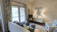 This two-floor space showcases a thoughtfully designed living area that combines comfort and contemporary style. The living room is anchored by a plush, light-toned sofa accented with an array of decorative pillows in grey and teal tones, complemented by tufted ottomans that add a touch of sophistication. Dark hardwood flooring grounds the space, providing a rich contrast to the neutral warm-beige walls.

A large sliding glass door floods the room with natural light while offering direct access to an outdoor area, framed by elegant patterned drapery panels that add visual interest. Blinds provide adjustable privacy and light control. A woven accent chair in dark tones occupies a corner beside decorative wall-mounted panels, creating a cozy reading nook.

Along one wall, a sleek white credenza with multiple drawers serves as both functional storage and a display surface, topped with matching ceramic table lamps and framed botanical artwork. The overall ambiance is calm and cohesive, with a carefully curated neutral palette punctuated by teal and grey accents that lend the space a polished, welcoming character.