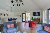 Located at 291 N Colonial Homes Circle NW in Atlanta, Georgia, this single-floor space combines modern design with a lively, functional layout suited for gathering and socializing. The interior is defined by soaring vaulted ceilings that amplify the sense of openness throughout, while light wood flooring runs continuously underfoot, lending warmth to the bright, white-walled interior.

The main lounge area features a seating arrangement of contemporary gray armchairs positioned around a bold abstract area rug, accented by a sputnik-style chandelier and a large picture window overlooking the surrounding neighborhood. Adjacent to this is a dining area anchored by a dark round table surrounded by striking red sculptural chairs, paired with a mid-century modern credenza and a wall-mounted flat-screen television.

A built-in white counter with blue modern barstools provides a secondary workspace or casual seating area, illuminated by industrial pendant lights and accented by colorful abstract artwork. Restroom access is conveniently located nearby.

Large black-framed glass doors and windows along the perimeter flood the space with natural light and open directly onto an outdoor deck overlooking a pool, seamlessly connecting the interior to the exterior environment. A ceiling fan provides additional comfort throughout.