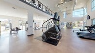 This two-story fitness center is a well-appointed facility designed to support a comprehensive workout experience. The main floor boasts soaring double-height ceilings with large clerestory windows that flood the space with natural light, creating an open and energizing atmosphere. A prominent ceiling fan complements the exposed ductwork above, adding an industrial yet modern aesthetic to the bright, white interior. The polished concrete flooring extends throughout, providing a durable and clean surface underfoot.

The ground level is generously equipped with a variety of cardio machines, including StairMaster climbers and rowing machines, as well as a full complement of strength-training equipment such as a multi-functional Smith machine with an adjustable bench, cable machines, and a range of selectorized weight stations. Wall-mounted televisions are positioned throughout for entertainment during workouts. Large glass doors and windows on multiple sides offer views of the surrounding outdoor areas, including a pool and landscaped grounds.

A staircase with wooden treads leads to the mezzanine level, which overlooks the main floor and houses additional cardio equipment, maximizing the facility's capacity. The upper level benefits from the same abundant natural light and open sightlines. The overall design is modern, clean, and functional, making this fitness center a standout amenity.