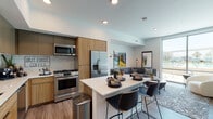 This thoughtfully designed single-floor residence showcases a contemporary aesthetic defined by clean lines, warm wood tones, and high-quality finishes throughout. The open-concept layout seamlessly connects the kitchen, dining, and living areas, creating a cohesive and functional living space.

The kitchen is a standout feature, equipped with stainless steel appliances including a gas range, built-in microwave, French door refrigerator, and dishwasher. A central island with a white quartz countertop provides additional prep space and casual seating for multiple bar-height chairs. Warm wood cabinetry is complemented by two-toned upper cabinets and a classic subway tile backsplash, adding texture and depth to the space. Wood-style flooring runs throughout, enhancing the cohesive feel.

The living area is bathed in natural light from expansive floor-to-ceiling windows and a sliding glass door, framing direct views of an outdoor pool and surrounding cityscape. The bedroom offers a calm retreat with neutral tones, a ceiling fan, and hardwood-style flooring.

The bathroom features large-format tile walls, a frameless glass walk-in shower, and wood-accented vanity cabinetry. A dedicated workspace nook visible from the kitchen adds versatile functionality to the overall layout.