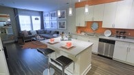 This single-floor residence showcases an open-concept layout that connects the kitchen and living area in a cohesive, modern design. The kitchen is a standout feature, centered around a spacious granite-topped island with seating for two sleek bar stools — ideal for casual dining or entertaining. White cabinetry lines the perimeter, complemented by a bold orange accent wall that adds a vibrant pop of color to the space. A stainless steel sink, dishwasher, and generous counter space complete the functional kitchen setup. Three contemporary pendant lights hang above the island, providing focused task lighting while enhancing the modern aesthetic. The living area flows naturally from the kitchen, featuring wood-look flooring, a large gray sectional sofa, and a gallery wall of framed artwork that adds character and depth. A window allows natural light to filter into the living space, brightening the overall ambiance. The consistent use of neutral tones accented with orange creates a cohesive and stylish atmosphere throughout, making this space both visually dynamic and highly functional for everyday living.