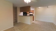 This single-floor residence features an open-concept design that seamlessly connects the kitchen, dining, and living areas, creating a cohesive and functional living environment. The kitchen is equipped with black appliances, including a range and overhead microwave, and is anchored by a peninsula with dark granite-style countertops that doubles as a casual dining or prep surface. Medium-toned wood cabinetry provides ample storage, while a large overhead cabinet structure adds a distinctive architectural element above the peninsula. A ceiling fan with a light fixture illuminates the main living area, which is finished with neutral wall-to-wall carpeting that extends throughout the space, lending warmth underfoot. To the left of the main living area, a built-in shelving unit offers practical display and storage opportunities, adding a custom feel to the interior. The walls are finished in a soft, neutral tone with white chair rail detailing, contributing to a clean and understated aesthetic. An adjacent room, visible through an open archway, provides additional living or dining space, further enhancing the versatility of the layout. Overall, the space presents a practical and comfortable interior with cohesive finishes.