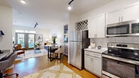 This single-floor residence offers a thoughtfully designed open-concept layout that seamlessly connects the kitchen, dining, and living areas. The kitchen is a standout feature, outfitted with gray shaker-style cabinetry, granite countertops, a classic white subway tile backsplash, a built-in wine rack, a stainless steel farmhouse sink, and a full suite of stainless steel appliances including a refrigerator, electric range, microwave, and dishwasher. Track lighting illuminates both the kitchen and the living area, where soaring ceilings and large windows invite abundant natural light. Warm hardwood-style flooring flows consistently throughout the main living spaces. The bedroom is a comfortable retreat, featuring elevated ceilings, a ceiling fan, and a generously sized window with greenery views. It connects directly to a full bathroom, finished with a granite-topped vanity, gray cabinetry, an illuminated mirror, and a tiled shower-tub combination. A spacious walk-in closet with multi-tiered wire shelving provides ample storage. The overall ambiance of the residence is clean, contemporary, and inviting, with consistent neutral tones and modern fixtures that create a cohesive and functional living environment.