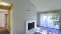 This single-floor space offers a clean, understated aesthetic defined by neutral tones and thoughtful architectural details. The living area serves as the heart of the space, drawing immediate attention with its soaring vaulted ceiling that adds a sense of openness and grandeur. A white brick fireplace anchors the room, providing a classic focal point that adds both character and warmth to the interior. The textured ceiling complements the space's design, adding a subtle layer of visual interest overhead. Dark hardwood-style flooring runs throughout, offering a rich contrast to the light gray walls and white trim. Natural light pours generously into the room through a large sliding glass door dressed with vertical blinds, casting bright, dynamic light across the floor and walls. The sliding door hints at outdoor access, enhancing the sense of connectivity to the exterior. A ceiling vent is thoughtfully integrated into the wall near the roofline, ensuring functional climate control without disrupting the room's clean lines. Overall, the space presents a bright, airy, and welcoming atmosphere, blending practical features with timeless design elements suited for comfortable everyday living.
