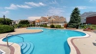 This outdoor amenity space centers around a generously sized freeform swimming pool, its crystal-clear turquoise water inviting relaxation under open skies. The pool's curvilinear design creates a resort-like atmosphere, complete with wide, gently stepped entry points that ease swimmers into the water. A broad concrete deck surrounds the pool, offering ample space for lounge chairs and casual outdoor furniture positioned to take full advantage of the sunny exposure.

Lush landscaping frames the area, with mature evergreen trees, ornamental shrubs, and well-maintained greenery providing a natural, park-like setting. Classic lamp posts punctuate the deck, adding a touch of traditional charm while ensuring the space remains functional into the evening hours.

At the rear of the pool area stands an attractive single-story clubhouse, designed in a traditional architectural style featuring a warm neutral siding, a pitched roof with dormers, a gazebo-style covered entrance, and a chimney. The structure adds a focal point to the landscape and likely serves as a community gathering space. Together, the pool, deck, landscaping, and clubhouse create a cohesive and welcoming outdoor environment ideal for leisure and recreation.