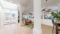 Located at 801 E Alosta Ave in Azusa, California, this thoughtfully designed amenity space offers a welcoming and functional environment across a single floor. The open main area greets visitors with soaring vaulted ceilings, light hardwood flooring, and an abundance of natural light streaming through large black-framed windows. Striking artistic murals and curated wall art lend the space a lively, creative energy, while a sculptural woven pendant light serves as a standout focal point overhead. Comfortable mid-century modern seating, including warm-toned accent chairs arranged around a wooden coffee table atop a green patterned rug, creates an inviting lounge atmosphere. A built-in counter with bar seating offers an alternative workspace within the open area. A hallway connects the main lounge to three private meeting rooms, each finished with sage green accent walls, textured carpet flooring, and light wood conference tables surrounded by cognac leather chairs. Each meeting room features its own framed artwork and is brightened by windows overlooking the surrounding landscape. One room includes a dedicated desk workspace, while all three offer a calm, productive setting ideal for focused work or small group collaboration.