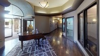 Located at 636 E Magnesium Rd in Spokane, Washington, this refined single-floor property offers a thoughtfully designed layout with upscale finishes and a warm, sophisticated ambiance throughout. The welcoming entry foyer sets an immediate tone of elegance, featuring rich hardwood flooring, a coffered tray ceiling with ambient lighting, a grand piano, and a decorative area rug — creating a striking first impression. Arched doorways and architectural detailing carry through the space, lending a cohesive and polished character. The main living area boasts soaring vaulted ceilings with a prominent arched window that invites abundant natural light, complemented by a tiled fireplace and carefully arranged seating. A secondary lounge area provides an intimate setting with additional vaulted ceilings and arched windows overlooking the outdoors. The kitchen is well-appointed with dark cabinetry, stone countertops, stainless steel appliances, a mosaic tile backsplash, and a breakfast bar with seating. A formal dining area with large windows offers a quiet, functional space. Additional rooms include a reception area displaying floor plans and branded signage, as well as a cozy sitting room with natural light and refined furnishings. Throughout, dark carpet and hardwood flooring, neutral wall tones, and curated décor create a consistently upscale and professional environment.