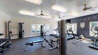 Located in Oak Grove, Houston, TX 77070, this dedicated fitness center offers a well-equipped, single-floor facility designed to support a comprehensive workout experience. The space is finished with durable carpet flooring throughout and features a clean, modern aesthetic with neutral gray walls accented by a bold dark feature wall bearing the word "FITNESS" in large lettering, adding a motivational focal point to the room.

Natural light pours in through numerous white-framed windows along multiple walls, creating a bright and energizing atmosphere. Ceiling fans provide additional ventilation and comfort during workouts, complemented by overhead fluorescent lighting for consistent illumination throughout the day and evening.

The cardio zone is well-appointed with several Star Trac treadmills and upright elliptical machines, all positioned to take advantage of the natural light and outdoor views. A wall-mounted flat-screen television serves the cardio area for entertainment. The strength training section includes a multi-station cable machine, a weight bench, and a well-stocked dumbbell rack offering a range of free weights. Additional functional fitness equipment includes a punching bag with stand, resistance bands, medicine balls, and exercise mats, supporting a variety of training styles. A pass-through window connects to an adjacent space, adding practical convenience to this fully outfitted community fitness facility.