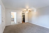 This single-floor unit offers a straightforward, functional layout designed for efficient use of space. The living area is finished with wall-to-wall carpet in a neutral tone and features a ceiling fan with integrated lighting, providing both comfort and airflow. Soft gray walls create a clean, understated backdrop, while a textured ceiling adds subtle dimension. An adjacent doorway connects the living space to the kitchen, where the flooring transitions to warm hardwood. The galley-style kitchen is equipped with a range and dishwasher on opposing sides, complemented by light-toned countertops and upper cabinetry, making efficient use of the available workspace. A black refrigerator anchors the kitchen's entrance. The bedroom is a well-proportioned, carpeted room with white walls, a window fitted with horizontal blinds that allows natural light to filter in, and a textured ceiling consistent with the rest of the unit. The entry area also features a window with blinds alongside a solid front door, offering a modest sense of arrival. Overall, the unit maintains a clean, neutral palette throughout, providing a blank canvas well-suited for personalization.