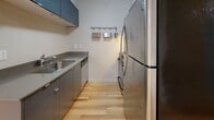 Located at 844 NE 69th St in Seattle's Roosevelt neighborhood, this one-bedroom, one-bathroom unit offers a well-organized layout across a single floor. The open living and dining area is a standout feature, showcasing warm hardwood flooring, recessed lighting, and multiple windows fitted with blinds that invite natural light while maintaining privacy. The kitchen is thoughtfully designed with sleek slate-blue cabinetry, stainless steel appliances including a full-size refrigerator and dishwasher, and a galley-style layout that maximizes functionality within a compact footprint. A hallway connects the main living area to the private quarters, featuring a wall-mounted coat rack for added convenience. The bedroom is carpeted and comfortably proportioned, offering a large window with views of surrounding greenery and direct access to the bathroom. The bathroom includes a full tub-shower combination with white tile surround, open shelving, and a vanity with matching cabinetry. A dedicated laundry area with a stacked washer and dryer adds practical value. Two closets provide ample storage throughout the unit.