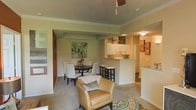 This single-story residence is designed with an open-concept layout that seamlessly connects the living, dining, and kitchen areas into one cohesive space. Crown moulding runs throughout, adding a classic architectural detail that enhances the overall aesthetic. The living area is carpeted and anchored by a decorative area rug, providing a warm and comfortable foundation for seating. A cushioned armchair in tan leather adds a relaxed yet refined touch to the space.

The dining area accommodates a round table surrounded by upholstered chairs, positioned beneath wall art that introduces soft botanical tones. This area flows naturally into the kitchen, which features a breakfast bar with bar stools, offering additional casual seating. The kitchen is outfitted with white cabinetry and light countertops, contributing to a clean and bright workspace. A peninsula counter separates the kitchen from the dining space while maintaining the open feel.

The warm beige and green color palette is consistent throughout, creating a cohesive and inviting ambiance. Ceiling-mounted lighting fixtures provide even illumination, complementing the overall welcoming and functional character of the home.