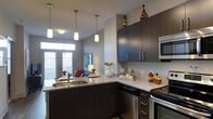 This single-floor residence offers a thoughtfully designed open-concept layout that seamlessly connects the kitchen and living areas, creating a cohesive and inviting atmosphere throughout. The kitchen serves as the centerpiece of the space, showcasing dark espresso cabinetry that extends to the upper walls for generous storage. Light-toned granite countertops provide a striking contrast, running along both the perimeter and a peninsula that doubles as a functional workspace and casual dining area. Three pendant lights hang above the peninsula, casting warm, focused illumination over the surface below. Stainless steel appliances, including a built-in microwave and dishwasher, lend the kitchen a polished, contemporary feel. The open peninsula overlooks the living area, which benefits from large windows and glass doors that flood the space with natural light and offer views to the outdoors. Neutral gray walls throughout create a calm, versatile backdrop that complements the darker cabinetry and light countertop tones. The overall ambiance is modern and clean, with a balance of dark and light elements that give the space a sophisticated yet comfortable character well-suited for everyday living and entertaining.