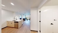 This well-designed 2-bedroom, 2-bathroom unit in Seattle's University District offers a thoughtfully efficient single-floor layout with modern finishes throughout. The open-concept living and kitchen area forms the heart of the space, featuring light wood-tone flooring that flows seamlessly from room to room. The kitchen is equipped with stainless steel appliances — including a full-size refrigerator, electric range, and microwave — complemented by light wood cabinetry, white quartz countertops, and a peninsula island with an undermount sink and striking matte black faucet. Pendant lighting above the island adds a polished touch, while bar seating makes the space ideal for casual dining. The living area is bright and airy, anchored by large windows that frame outdoor views of the surrounding neighborhood and feature a wall-mounted split AC/heating unit for year-round comfort. Each bedroom is a well-proportioned retreat with built-in storage and ceiling fans, while the primary bedroom benefits from a window with scenic views. Both bathrooms mirror each other in design, each featuring a tub-shower combination enclosed by sliding glass doors with matte black frames, light wood vanities, and wood-look tile flooring — offering a clean, cohesive aesthetic throughout.