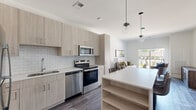 Located at 250 Plaza Circle in Mundelein, IL, this well-appointed 3-bedroom, 2-bathroom residence offers a thoughtfully designed single-floor layout with modern finishes throughout. The open-concept kitchen and living area serves as the heart of the home, featuring light wood cabinetry, a white subway tile backsplash, stainless steel appliances, and a large kitchen island with built-in shelving and seating — ideal for both meal prep and casual dining. Globe pendant lights add a contemporary touch overhead. The living room is bright and airy, with wide-plank hardwood-style flooring, white walls, and a sliding glass door that opens to a private balcony with outdoor views. The spacious primary bedroom is carpeted and filled with natural light from a large window overlooking the exterior. Two full bathrooms include a glass-enclosed walk-in shower with textured tile surround. A dedicated closet features wire shelving and hanging rods for ample storage. The unit also includes in-unit laundry. Soft carpet in the bedrooms and hallways contrasts with the sleek hard flooring in the main living areas, creating a comfortable and cohesive living environment throughout.