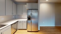 Located at 72 N Clinton Ave in the heart of downtown Rochester, NY, this single-floor unit presents a clean, modern aesthetic defined by neutral tones, hardwood-style flooring, and an efficient layout. The open-concept living and kitchen area forms the core of the space, where warm wood flooring flows seamlessly from the main living zone into the kitchen. Large windows fitted with roller shades allow natural light to fill the living area, offering views of the surrounding urban streetscape. Track lighting overhead provides additional illumination throughout.

The kitchen is thoughtfully designed with gray flat-front cabinetry extending from floor to ceiling, offering generous storage. Stainless steel appliances—including a side-by-side refrigerator with ice and water dispenser, electric range, over-the-range microwave, and dishwasher—complement the white countertops and tile backsplash. Light wood flooring continues underfoot, maintaining visual continuity.

A private bedroom provides a calm retreat, featuring neutral wall tones, a window with a roller shade, hardwood-style flooring, and a closet with a paneled white door. The bathroom is clean and functional, finished with the same wood-tone flooring, neutral gray walls, and a full tub-shower combo with chrome fixtures.