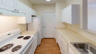 This single-floor unit offers a thoughtfully arranged layout with distinct living, kitchen, bedroom, and bathroom spaces. The living area features soft neutral-toned carpet, crown molding, a ceiling fan with light fixture, and a window overlooking an outdoor balcony, creating a bright and comfortable atmosphere.

The galley-style kitchen is efficiently designed with white cabinetry lining both walls, light countertops, wood-look flooring, and white appliances including a coil-burner range, dishwasher, and refrigerator. A pass-through counter connects the kitchen to the living area, enhancing the open feel of the shared space.

The bedrooms are carpeted and finished in warm neutral tones. One bedroom features a charming bay window configuration that draws in generous natural light and offers exterior views of the surrounding neighborhood. A second bedroom includes a standard window and textured ceiling.

The bathroom is well-equipped with a vanity featuring an integrated sink and mirror, wood-look flooring, and a full-size soaking tub and shower combination accented by a distinctive checkerboard tile border in muted earth tones. Throughout the unit, clean white trim, consistent neutral walls, and ample natural light contribute to an overall bright and cohesive aesthetic.