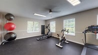 Located at 701 Baywood Cir in Morrow, Georgia, this single-story fitness center offers a spacious and well-organized workout environment designed to accommodate a variety of exercise routines. The facility is laid out across an open, L-shaped floor plan, providing ample room for both cardio and strength training activities without feeling crowded.

The flooring throughout is durable rubber gym matting in a dark speckled finish, well-suited for high-traffic fitness use. Neutral beige walls are accented by a continuous chair rail detail, lending a clean and polished appearance to the space. Textured ceilings are fitted with flush fluorescent light fixtures and ceiling fans, ensuring consistent lighting and air circulation throughout.

Multiple windows fitted with white horizontal blinds line the walls, drawing in generous natural light and offering views of the surrounding greenery. The cardio zone is equipped with multiple treadmills and an elliptical trainer, while a stationary spin bike and rowing machine provide additional low-impact options. The strength training area includes a cable machine, a weight bench, a dumbbell rack with a full set of free weights, and additional resistance equipment. The facility connects to an adjacent room, adding further functional versatility to the overall space.