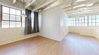 Located in Oakland's Fruitvale neighborhood, this two-story live-work loft blends industrial character with functional residential living. The lower level features a vast, open-plan living area with polished concrete floors, an exposed beam and joist ceiling, and abundant natural light streaming through large factory-style grid windows. A staircase leads to the upper level, where warm laminate hardwood flooring runs throughout. The kitchen is equipped with oak cabinetry, dark countertops, a stainless steel gas range, and a full-size refrigerator, complementing an open dining area. The living space on the upper floor continues the industrial aesthetic with striking white-painted exposed beam ceilings and generous windows that fill the space with natural light. The bedroom is a comfortable, well-lit room featuring grid-pane windows and the same warm hardwood flooring. The full bathroom includes a shower-tub combination and hardwood-style flooring. Throughout both floors, white walls and open structural ceilings create an airy, creative ambiance ideal for live-work arrangements. The generous square footage across two levels provides ample room for both living and working in this distinctive Oakland loft.