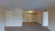 This single-floor space presents a straightforward and functional layout centered around a generously sized main room. The room features wall-to-wall carpeting in a neutral beige tone, providing a soft and comfortable underfoot feel throughout the space. The walls are finished in a light, neutral color palette that contributes to an open and airy ambiance, while classic crown molding along the ceiling adds a subtle architectural detail that elevates the overall finish of the room. A flush-mount ceiling light provides centralized overhead illumination, ensuring the space is well-lit. A ceiling-mounted HVAC vent is also present, indicating integrated climate control for year-round comfort. White baseboards frame the perimeter of the room, offering a clean contrast against the carpeted floor. A doorway with a white-trimmed surround leads to an adjacent area, hinting at additional connected spaces within the layout. A fire extinguisher is mounted near the doorway, reflecting attention to safety standards. The overall atmosphere is calm and neutral, making the space highly adaptable for a variety of uses and arrangements.
