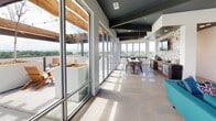 This rooftop amenity space is a thoughtfully designed retreat that seamlessly blends indoor comfort with outdoor living. The interior lounge area features a dramatic dark geometric ceiling with recessed lighting, crisp white walls, and large floor-to-ceiling windows that flood the space with natural light. Vibrant teal seating, a leather accent chair, a wall-mounted display, and a media credenza create a welcoming, modern atmosphere ideal for gathering.

Adjacent to the lounge, a full kitchen and dining area runs along the perimeter, finished with polished tile flooring, warm wood ceiling beams, dark cabinetry, and stainless steel appliances. A long communal dining table anchors the space, while floor-to-ceiling sliding glass doors along one entire wall open directly onto the outdoor terrace, allowing for an effortless indoor-outdoor flow.

The outdoor terraces are generously proportioned and feature multiple seating arrangements, including wicker lounge sets with cushions, vibrant green accent chairs, and teak Adirondack chairs positioned beside large raised planters. A wood pergola structure with string lights adds warmth and character overhead. Sweeping panoramic views of the surrounding cityscape and tree canopy extend in multiple directions, making the outdoor spaces a standout feature of this elevated, amenity-rich environment.