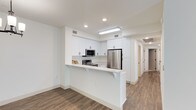 This well-appointed single-floor space showcases a clean, modern aesthetic defined by crisp white walls, warm wood-look flooring, and thoughtful design throughout. The kitchen is a standout feature, boasting an abundance of white shaker-style cabinetry that extends to the ceiling, providing generous storage. Stainless steel appliances — including a refrigerator, microwave, and range — lend a polished, contemporary feel. A white peninsula with corbel bracket supports doubles as a breakfast bar, creating a natural divide between the kitchen and the adjoining living area while offering a casual dining or workspace surface. Recessed lighting and a flush-mount ceiling fixture ensure the kitchen is well-lit, complementing the bright, airy atmosphere. The wood-look flooring runs seamlessly throughout the space, adding warmth and continuity. A hallway visible in the background leads to additional rooms, suggesting a practical and efficient layout. The overall ambiance is fresh, clean, and move-in ready, making it an inviting and functional living environment.
