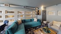 Located at 2005 W Campbell Rd in Garland, Texas, this single-floor amenity lounge is a thoughtfully designed entertainment and social space. The interior is anchored by a bold blue-and-white abstract patterned carpet that flows throughout, lending the space a cohesive, contemporary feel. Exposed metallic ductwork runs along the upper portions of the walls and ceiling, adding an industrial edge that contrasts with the otherwise polished décor.

The space is divided into distinct functional zones. A media lounge area features a wall-mounted flat-screen television above a wood console, flanked by decorative wall art and recessed lighting. Adjacent to this is a dedicated gaming area equipped with multiple round poker-style tables surrounded by wood and leather chairs, with an additional wall-mounted TV for viewing. A dartboard cabinet is mounted nearby, enhancing the recreational atmosphere.

A separate seating nook features a large teal sectional sofa positioned beside a distinctive open-grid partition wall, providing visual separation while maintaining an open feel. A gold-accented round table with navy chairs nearby offers a casual spot for games or conversation.

A glass panel garage door opens to a balcony area, flooding the space with natural light and providing views of the surrounding exterior. A private entry door and built-in display shelving complete the well-appointed layout.