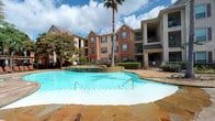 This residential community amenity space offers two distinct areas designed to support an active lifestyle. The outdoor area showcases a generously sized resort-style swimming pool with a freeform design, complemented by a raised spa with water features. The pool deck is finished with natural stone pavers in warm earth tones, creating an inviting atmosphere. Towering palm trees, mature landscaping, and well-maintained shrubbery frame the space, while the surrounding multi-story brick and neutral-tone buildings provide a sense of enclosure and privacy. The fitness center is a spacious, single-floor facility featuring soaring ceilings that lend an open, airy feel. The interior is finished with clean white walls and durable rubberized flooring suited for high-traffic athletic use. The facility is equipped with a comprehensive array of Life Fitness strength machines, cable systems, free weights, a weight bench, cardio equipment including treadmills and stationary bikes, and a tire sled for functional training. Full-length mirrors along one wall visually expand the space and support proper form during workouts. Wall-mounted televisions and recessed lighting enhance the workout environment. French door entry with glass panels allows natural light into the space, while an exercise reference chart and sanitizing station promote safe, informed use.