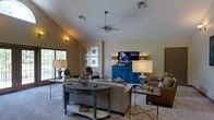 Located at 342 Riverwood Dr. in Crestview, Florida, this single-story home offers a thoughtfully designed layout with standout architectural details. The living area is the centerpiece of the home, showcasing soaring vaulted ceilings that create an expansive, airy atmosphere. A ceiling fan is centrally mounted, contributing both style and comfort to the space. Neutral wall tones provide a warm, welcoming backdrop that complements the room's furnishings and décor. Natural light filters in through windows along the wall, brightening the interior and adding to the open feel of the space. The living room is well-appointed, featuring a comfortable seating arrangement, glass-top accent tables, and a distinctive blue media console that adds a bold pop of color. A wall-mounted flat-screen television serves as a focal point, flanked by elegant table lamps. Framed artwork is displayed in a grid arrangement on the wall, adding a curated, decorative touch. The carpeted flooring adds warmth underfoot, tying the room together cohesively. Overall, this home combines practical single-floor living with inviting design elements, making it a comfortable and visually appealing residence in Crestview, FL.