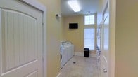 This single-floor shared laundry facility is designed with practicality and comfort in mind. The room is finished with warm yellow walls and neutral tile flooring, creating a clean and welcoming atmosphere. A row of full-sized washing machines and dryers lines one wall, providing ample capacity for residents. Opposite the machines, additional laundry equipment is accessible along the far wall, maximizing the room's utility. Natural light filters in through a high window fitted with horizontal blinds, complementing the bright overhead fluorescent lighting that keeps the space well-illuminated at all times. A wall-mounted flat-screen television is positioned for easy viewing, offering entertainment during wash cycles. Security is addressed through a ceiling-mounted surveillance camera, ensuring a safe environment. A waste bin is conveniently placed near the center of the room for easy access. The layout is thoughtfully organized with clear pathways between equipment, and a door on the left side of the room provides straightforward entry and exit. Overall, the space balances functional design with user comfort, making it a practical and pleasant shared amenity.