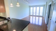 This single-floor space offers a seamless, open-plan layout that effortlessly connects the kitchen and living areas. Rich hardwood floors run throughout, reflecting natural light and adding warmth to the interior. The kitchen is anchored by a striking dark granite countertop, complemented by stylish pendant lighting that hangs above, creating a refined focal point at the heart of the space. Track lighting on the ceiling provides additional versatility for illuminating the area. The living space is bathed in natural light, thanks to a set of elegant French doors that stretch toward the ceiling and open onto a private balcony. The doors frame views of the surrounding cityscape, blending indoor comfort with an outdoor connection. Soft, neutral wall tones in a gentle sage-green hue lend a calm and airy ambiance throughout. Clean white baseboards and trim add a crisp, polished finish to the overall aesthetic. The open flow between the kitchen and living areas makes this space feel expansive and welcoming, ideal for both everyday living and entertaining. Every detail, from the gleaming floors to the abundance of natural light, contributes to a bright, modern, and inviting atmosphere.