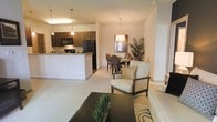 This single-floor residence offers an open-concept design that seamlessly integrates the kitchen, dining, and living areas into one cohesive and functional living space. The kitchen is equipped with dark cabinetry, black appliances including a refrigerator and microwave, and a breakfast bar countertop that serves as both a prep area and casual dining spot. Pendant lighting above the bar adds a warm, focused glow to the space. Adjacent to the kitchen, a dedicated dining area features a round table with upholstered chairs and a stylish overhead pendant fixture, making it ideal for everyday meals or intimate gatherings. The living area flows naturally from the dining space and is highlighted by neutral-toned furnishings, including a cushioned armchair, a sleek coffee table, and a plush area rug with a bold pattern. The overall color palette is soft and neutral, with warm beige walls and light flooring contributing to a calm and inviting ambiance throughout. Thoughtful accent decor, including potted greenery and a wall mirror in the dining area, adds depth and character. The layout maximizes the sense of openness, making the space feel airy and well-connected throughout.