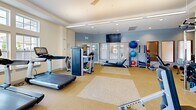 Located at 243 Steele Rd in West Hartford, CT, this fitness center offers a generously proportioned, single-floor exercise space designed to support a comprehensive workout experience. The room is bathed in natural light courtesy of large, multi-pane windows lining multiple walls, providing open views of the surrounding community and creating an energizing atmosphere throughout the day. Ceiling fans and recessed lighting complement the bright interior, maintaining comfort during exercise.

The space is thoughtfully organized to accommodate a diverse range of fitness activities. A robust row of treadmills lines one side of the room, accompanied by elliptical trainers and stationary bikes, all equipped with individual digital displays. Strength training is well represented with a dedicated multi-station cable machine and free weight equipment. For functional fitness and flexibility work, stability balls, resistance accessories, and additional benches are neatly arranged near the entrance area.

The flooring consists of durable, speckled rubberized material that provides cushioning and traction underfoot. Full-length mirrors are positioned strategically to aid in form monitoring during strength exercises. Wall-mounted flat-screen televisions are installed throughout the room, offering entertainment during cardio sessions. The overall design is clean, modern, and practical, making this fitness center a well-rounded facility for residents seeking convenient, on-site exercise amenities.