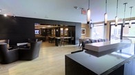 This single-floor space presents a contemporary open-concept layout designed for socializing and relaxation. The interior is defined by a sophisticated contrast of dark accent walls and warm, light-toned hardwood flooring, creating a polished yet inviting atmosphere. Multiple distinct seating zones are arranged throughout, including a lounge area with rounded upholstered chairs, a bar-height dining section with dark counter-style tables and matching stools, and additional casual seating near large windows that invite natural light into the space. A wall-mounted flat-screen television anchors one end of the room, enhancing the lounge-like character. Pendant lighting, including cylindrical fixtures and a globe-style accent light, adds refined detail overhead and contributes to the ambient warmth of the space. Framed artwork punctuates the walls, adding visual interest. A dark reception or service counter in the foreground suggests a staffed or managed element to the space. The overall design balances modern minimalism with functional comfort, making it well-suited as a shared amenity or hospitality lounge.