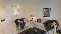 This single-story residence offers a comfortable and functional layout designed for everyday living. The living room serves as the heart of the home, featuring light-toned tile flooring that extends throughout the space, lending a clean and cohesive feel. Neutral white walls provide a bright, airy backdrop, while a ceiling fan overhead ensures year-round comfort. A notable architectural detail is the pass-through window that opens directly into the kitchen, creating a convenient connection between the two spaces and allowing natural light to flow through. The kitchen, visible through the pass-through, features cabinetry and appears well-appointed for its size. The living area provides ample room for seating arrangements, with space to accommodate multiple sofas and accent furniture, including side tables and a central coffee table. Framed wall art adds a decorative touch, contributing to the warm and inviting ambiance of the room. The overall atmosphere is understated and practical, with a layout that makes efficient use of space while maintaining a welcoming character throughout.