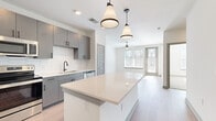 This well-designed single-floor unit offers a thoughtful layout that maximizes functionality across its comfortable footprint. The open-concept kitchen and living area serves as the heart of the space, where the kitchen stands out with gray flat-front cabinetry, a geometric white tile backsplash, quartz countertops, and a full suite of stainless steel appliances including a refrigerator with ice maker, electric range, microwave, and dishwasher. A central island with a white quartz surface provides additional prep space and casual seating. Stylish cone-shaped pendant lights hang above, complementing the recessed lighting throughout. Light hardwood-style flooring extends across the main living areas, contributing to a bright, cohesive aesthetic. The living room receives ample natural light through large windows overlooking a wooded outdoor view, and is equipped with a ceiling fan. The bedroom features plush carpeting, a ceiling fan, and a generous window that fills the room with natural light. An adjacent walk-in closet offers multi-level wire shelving for organized storage. The bathroom includes a soaking tub with tile surround, a floating gray vanity, and light wood-look flooring, maintaining the unit's clean, modern palette throughout.