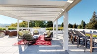This rooftop terrace is a thoughtfully designed outdoor amenity space that blends comfort, functionality, and scenic beauty. The centerpiece of the terrace is a large white pergola structure, strung with bistro lights, that provides partial shade over a generous lounge area furnished with woven rattan seating, accent chairs, and a bold red patterned area rug — creating an inviting social setting ideal for gatherings. Adjacent to the covered lounge, the open-air portion of the terrace extends into multiple distinct zones. A high-top bar dining table with counter stools offers an elevated dining experience, while separate dining tables with modern chairs provide additional seating options throughout. A fully equipped outdoor kitchen lines one side of the terrace, featuring multiple built-in stainless steel grills and prep surfaces — perfect for al fresco entertaining. Raised planters filled with ornamental grasses and lush greenery line the perimeter, adding a natural element and softening the clean, modern aesthetic. Large-format tile pavers cover the entire terrace floor, lending a polished, cohesive look. Panoramic views of the surrounding tree canopy and rolling hills are visible from all areas of the terrace, bathed in natural sunlight beneath a clear blue sky.