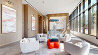 Astra Tower's ground-floor lobby is a masterfully designed, expansive space that establishes an immediate sense of refined elegance. The building's exterior presents a grand colonnade with tall white columns, a warm wood-clad soffit, and manicured planters that frame the entrance along the urban streetscape.

Upon entry, soaring floor-to-ceiling glass walls flood the interior with abundant natural light, offering open views of the surrounding cityscape. The main reception area is anchored by a striking marble-and-wood concierge desk beneath the illuminated "Astra Tower" signage, adorned with a cascading arrangement of sculptural floral wall art. A curated seating vignette featuring plush swivel chairs arranged on a circular area rug centers the welcoming lounge, accented by a vibrant red sculptural table and a glass display model of the tower itself.

Adjacent to the reception is a richly appointed library lounge, lined floor-to-ceiling with warm wood-toned bookshelves and accented by recessed lighting. A large-scale landscape artwork serves as a focal point above a solid wood bench, while a contemporary globe chandelier adds a sophisticated finishing touch. Neutral stone tile flooring runs seamlessly throughout, unifying the two distinct lounge areas into a cohesive, inviting environment that balances modern design with warmth and functionality.